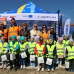 Group of smiling children in neon yellow vests posing with adults in orange safety jackets at an outdoor community event under a blue tent with logos in the background.