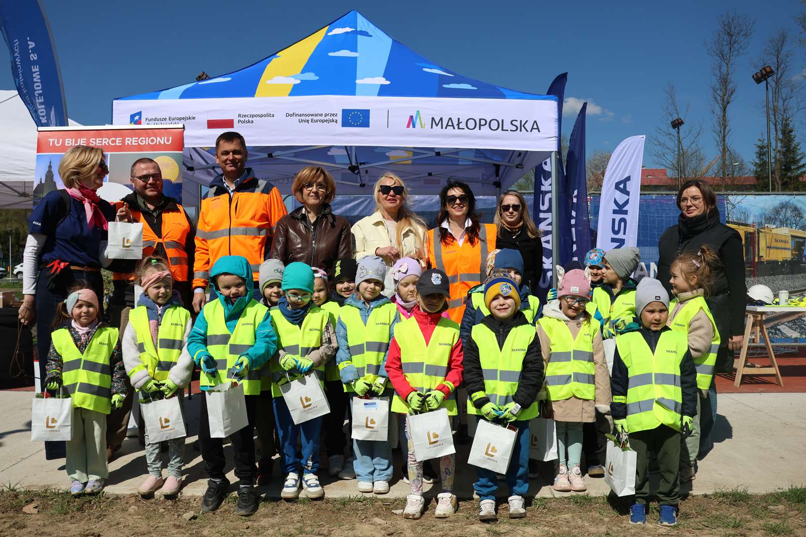 Group of smiling children in neon yellow vests posing with adults in orange safety jackets at an outdoor community event under a blue tent with logos in the background.