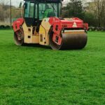 A tandem road roller and compactor on a grassy field, likely preparing ground for paving, with an operator in the cab.