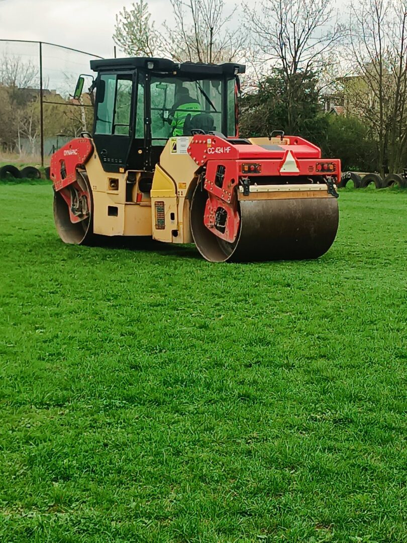A tandem road roller and compactor on a grassy field, likely preparing ground for paving, with an operator in the cab.