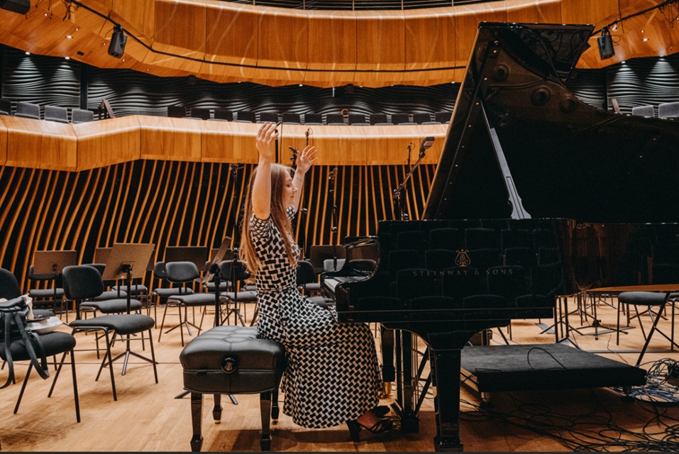 Woman in a black-and-white patterned dress raises arms while seated at a grand piano on a concert hall stage.