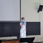 Girl in a light blue floral dress stands on a classroom stage, holding a blue handheld fan in front of her face.