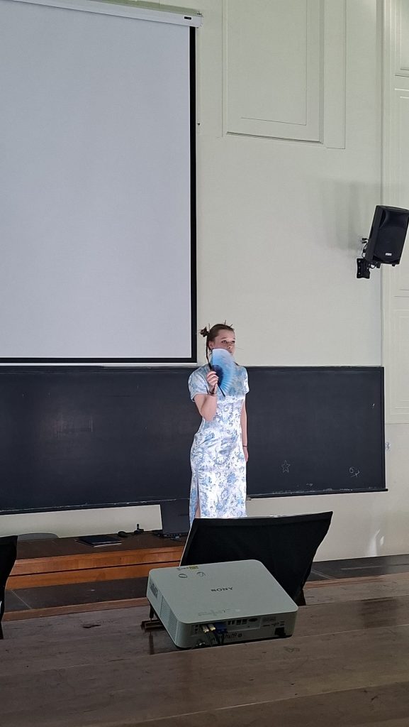 Girl in a light blue floral dress stands on a classroom stage, holding a blue handheld fan in front of her face.