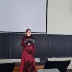 Person in a long red dress stands on a stage in a classroom, holding a black circular prop in front of a large blank projector screen and chalkboard backdrop.