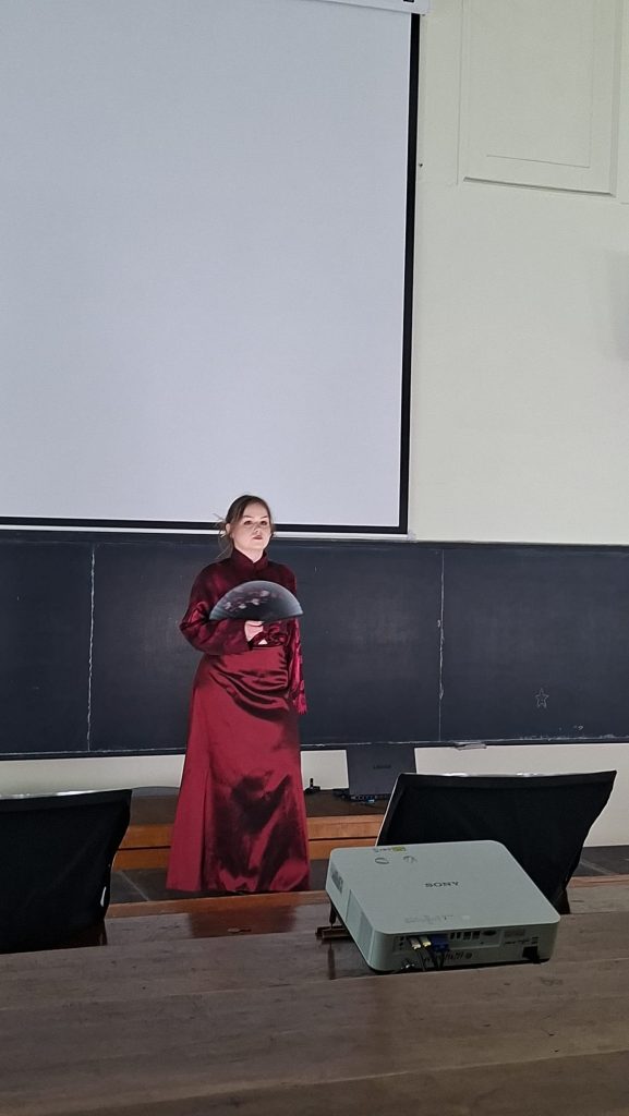 Person in a long red dress stands on a stage in a classroom, holding a black circular prop in front of a large blank projector screen and chalkboard backdrop.