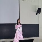 Woman in a light pink ball gown holding a pink handheld fan, standing in front of a large projection screen in a classroom or lecture hall.