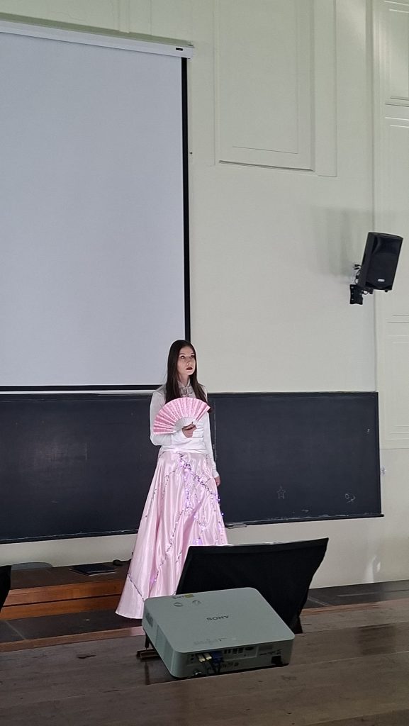 Woman in a light pink ball gown holding a pink handheld fan, standing in front of a large projection screen in a classroom or lecture hall.
