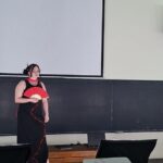 A performer in a black dress holds a red folding fan on a classroom stage in front of a projection screen, ready to perform.