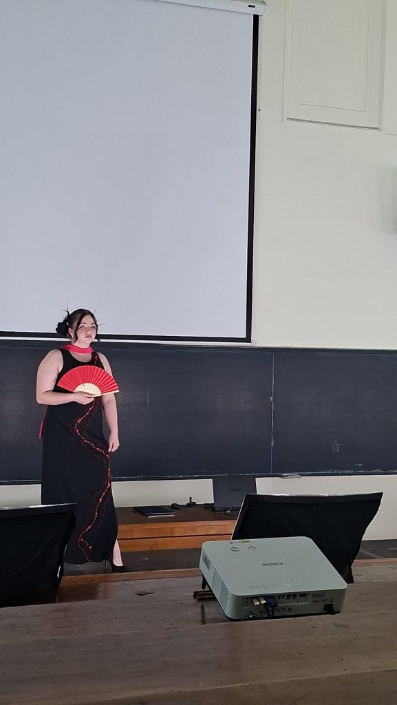 A performer in a black dress holds a red folding fan on a classroom stage in front of a projection screen, ready to perform.