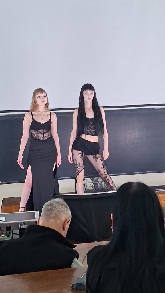 Two models in black lace outfits pose on a stage in front of a chalkboard as an audience watches a fashion show.