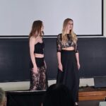 Two young women in black lace outfits stand on a stage in front of a large screen, facing each other, with audience silhouettes in the foreground.