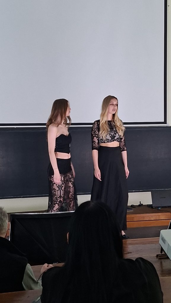 Two young women in black lace outfits stand on a stage in front of a large screen, facing each other, with audience silhouettes in the foreground.