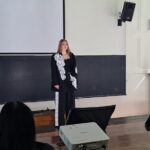 Woman in a black outfit with white lace stands on a stage in a lecture hall, presenting in front of a large projector screen and a speaker on the wall. Audience members sit at wooden benches in the foreground.