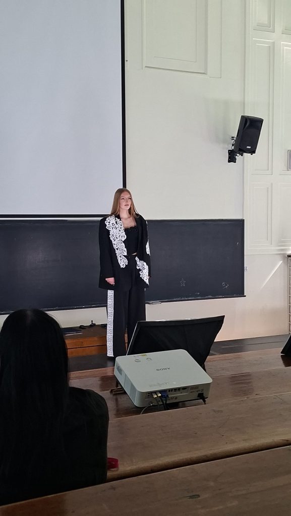 Woman in a black outfit with white lace stands on a stage in a lecture hall, presenting in front of a large projector screen and a speaker on the wall. Audience members sit at wooden benches in the foreground.