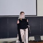 Person standing at front of a classroom/projection screen, wearing a black cold-shoulder top and white drawstring pants, with a light scarf over their face, addressing an audience.