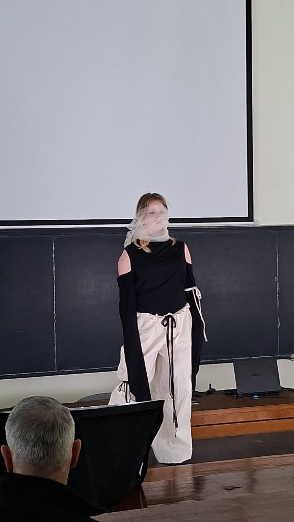 Person standing at front of a classroom/projection screen, wearing a black cold-shoulder top and white drawstring pants, with a light scarf over their face, addressing an audience.