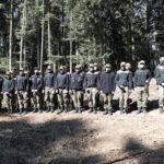A line of soldiers in black jackets and camouflage pants standing in a sunlit forest clearing.