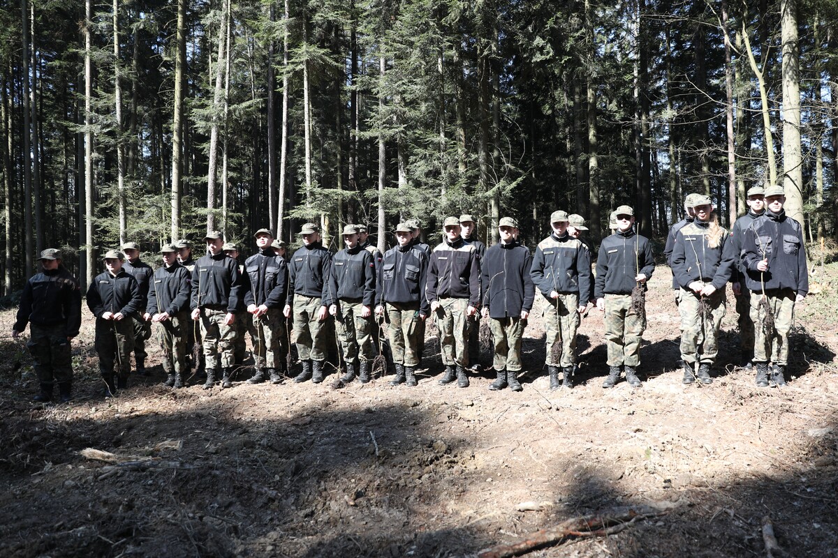 A line of soldiers in black jackets and camouflage pants standing in a sunlit forest clearing.
