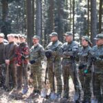 Line of soldiers and trainees in camouflage standing in a forest, each holding a shovel for outdoor work or training. (Informative)