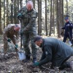 Soldiers and a civilian digging in a forest clearing, using shovels to excavate soil together.