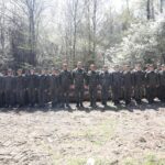 Line of soldiers in camouflage uniforms standing side by side outdoors in a forest, logs behind them.