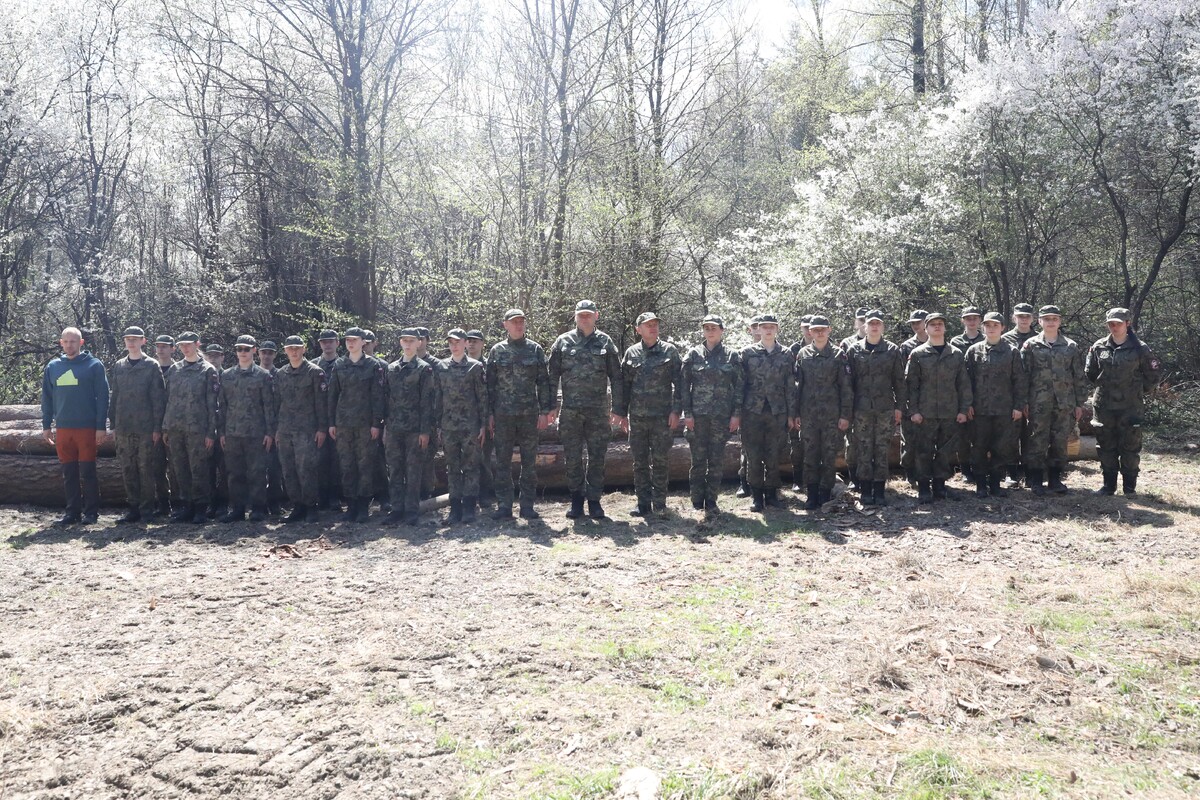 Line of soldiers in camouflage uniforms standing side by side outdoors in a forest, logs behind them.