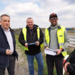 Four men standing on a roadside at a construction site, two wearing neon safety vests and holding blue folders, appearing to discuss plans.