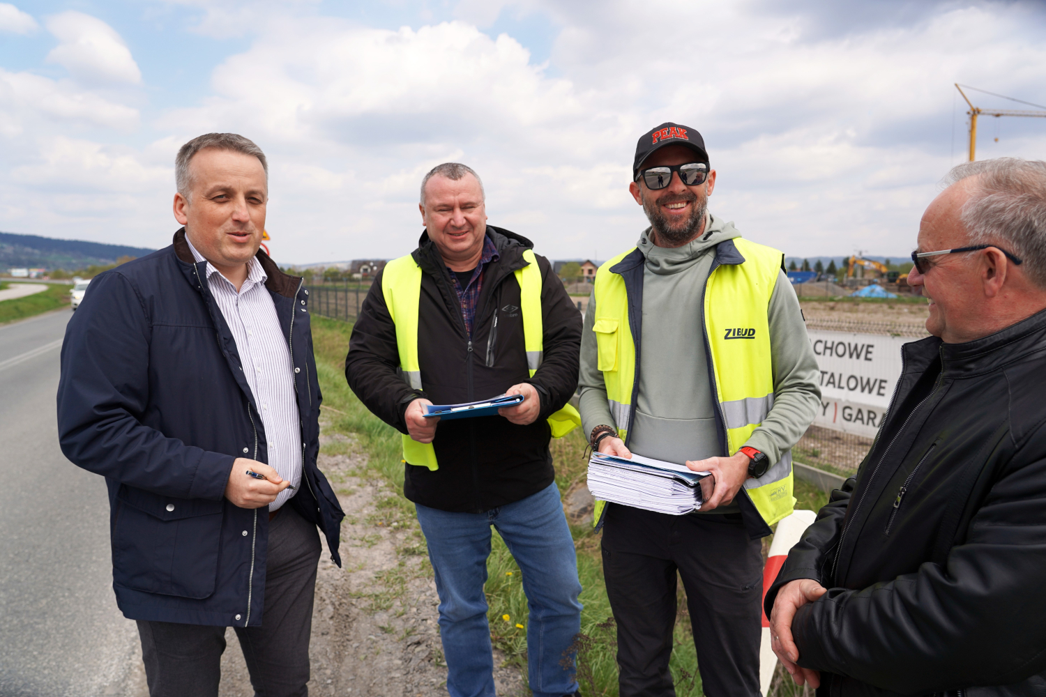 Four men standing on a roadside at a construction site, two wearing neon safety vests and holding blue folders, appearing to discuss plans.
