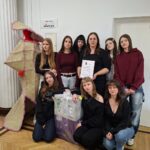 Group of nine young people posing for a photo indoors, one holding a diploma while another holds a large wrapped prize; a straw sculpture stands to the left and an eduroam sign is on the wall behind them.