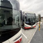 Several white and red electric buses parked at a paved loading area, with a charging station on the right and a cable connected to the unit.