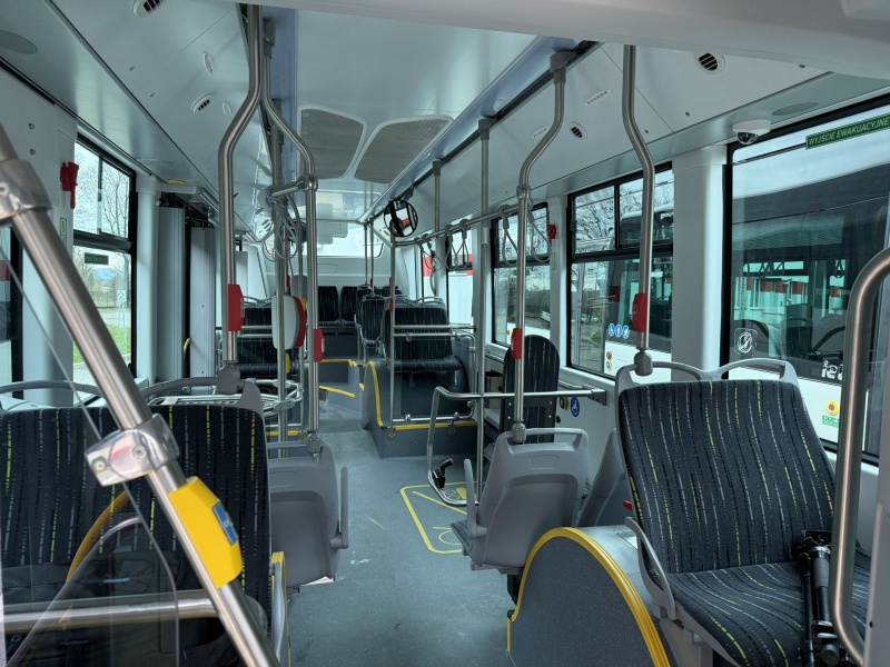 Interior of an empty city bus with rows of dark seats, metal handrails, and big windows; yellow priority seating and obvious aisle leading toward the front.