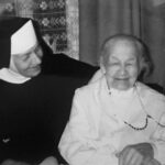 Nun in a black habit with white coif sits beside a smiling elderly woman in white clothing and a beaded necklace.