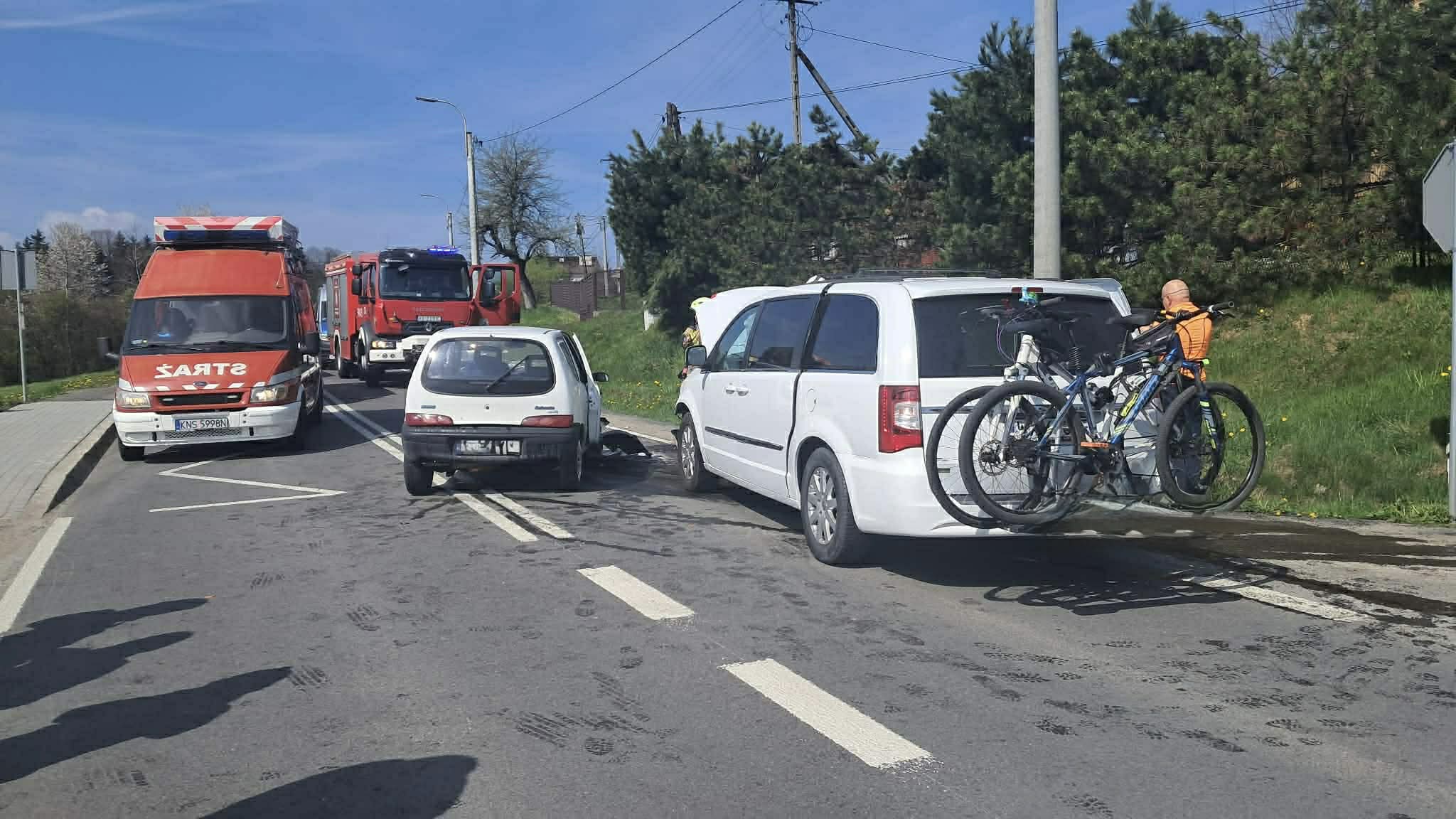 Emergency vehicles stop on a city road as a white van with bikes attached sits on the right; person in orange jacket nearby.