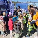 Group of children lined up outdoors while staff in high-visibility vests prepare to give them something at a Skanska event.