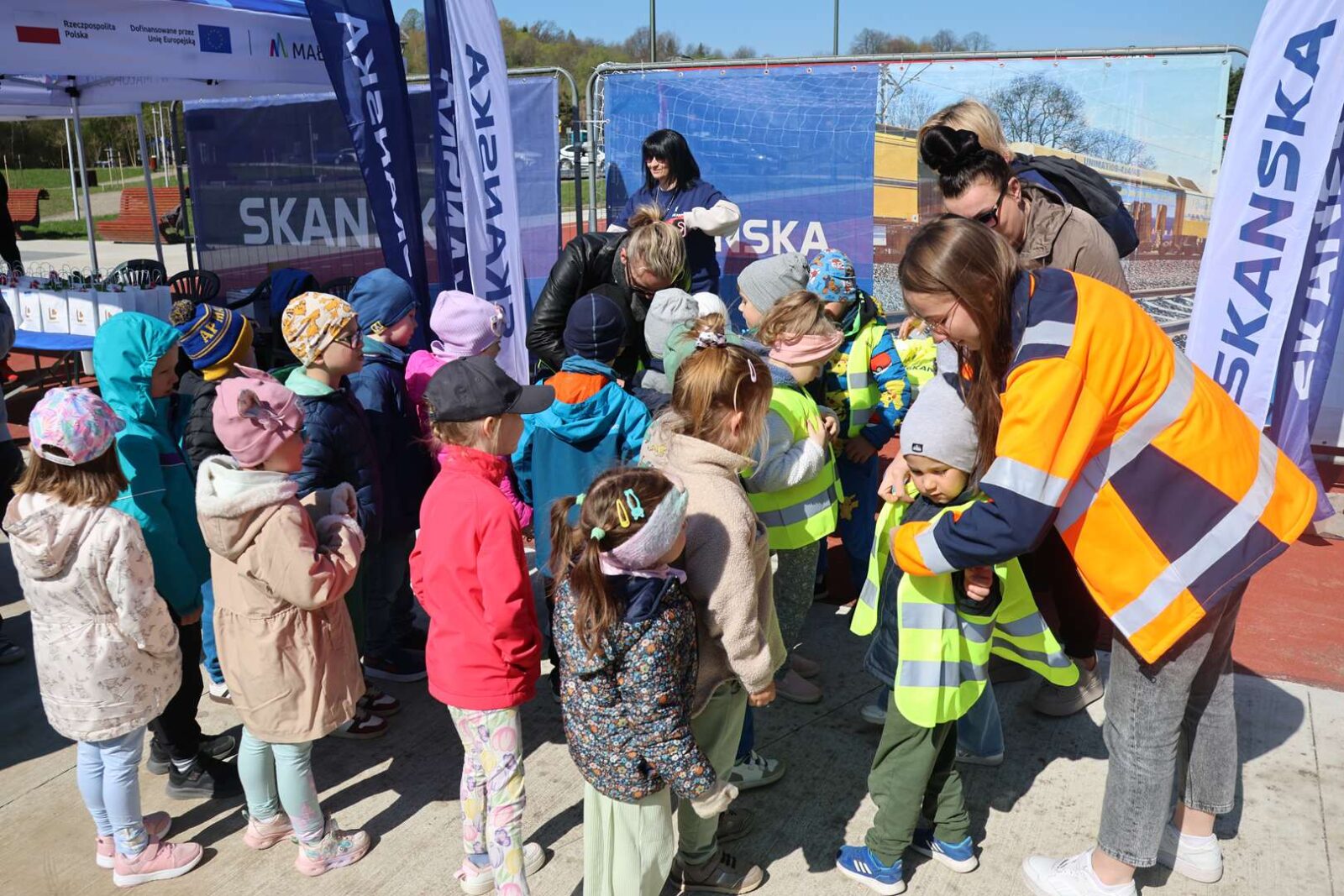 Group of children lined up outdoors while staff in high-visibility vests prepare to give them something at a Skanska event.