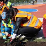 Children and adults in high-visibility vests planting flowers in a soil bed beside a running track on a sunny day; community gardening activity.
