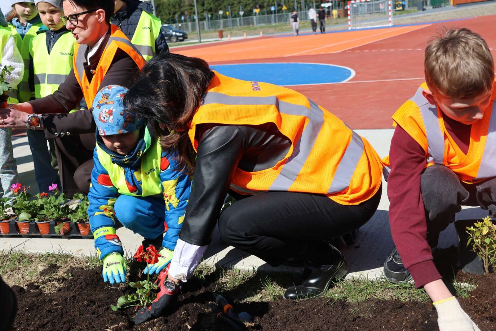 Children and adults in high-visibility vests planting flowers in a soil bed beside a running track on a sunny day; community gardening activity.