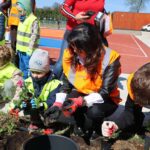 Children in neon safety vests plant flowers in a garden bed beside a running track on a sunny day, guided by adults.