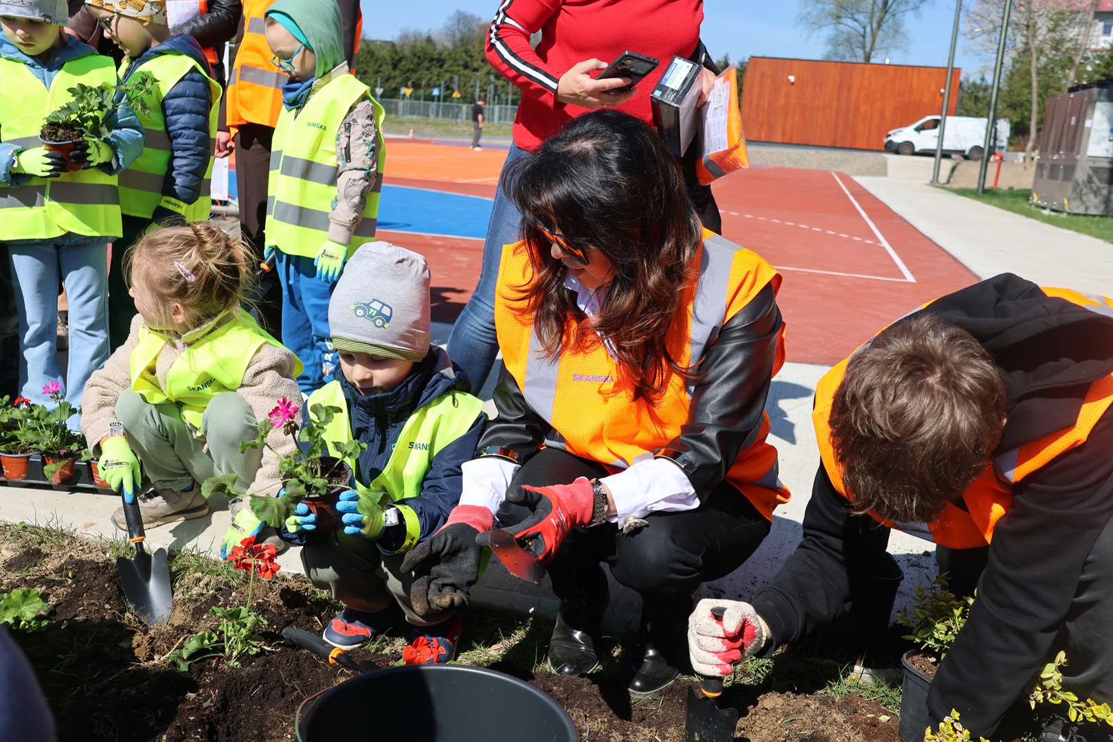 Children in neon safety vests plant flowers in a garden bed beside a running track on a sunny day, guided by adults.