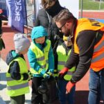 Instructor in a bright orange vest guiding kids in neon vests as they inspect a helmet on a track barriered by a running track setting.