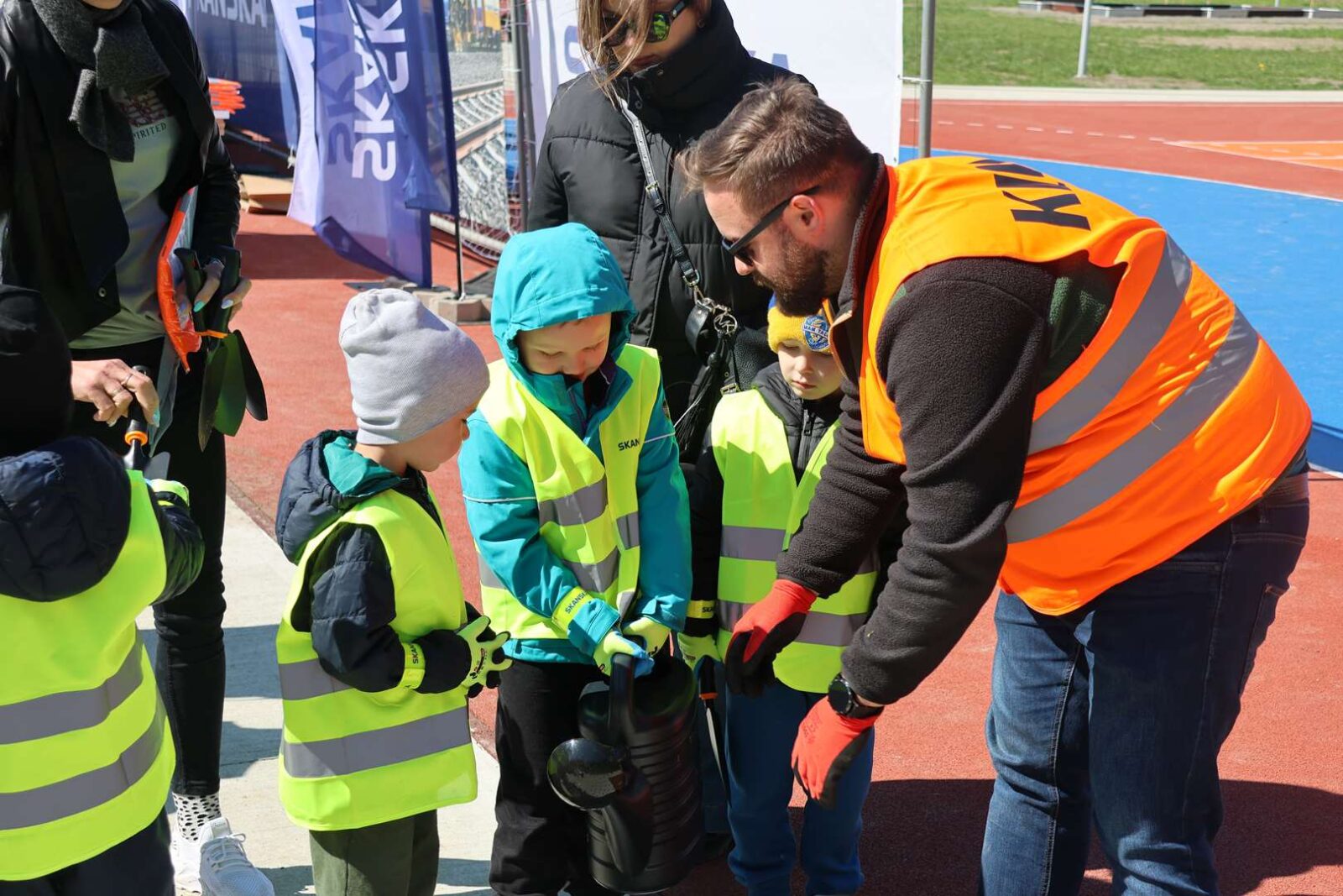 Instructor in a bright orange vest guiding kids in neon vests as they inspect a helmet on a track barriered by a running track setting.
