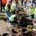 Group of kids in neon safety vests planting flowers in a garden bed under adult supervision outdoors.",