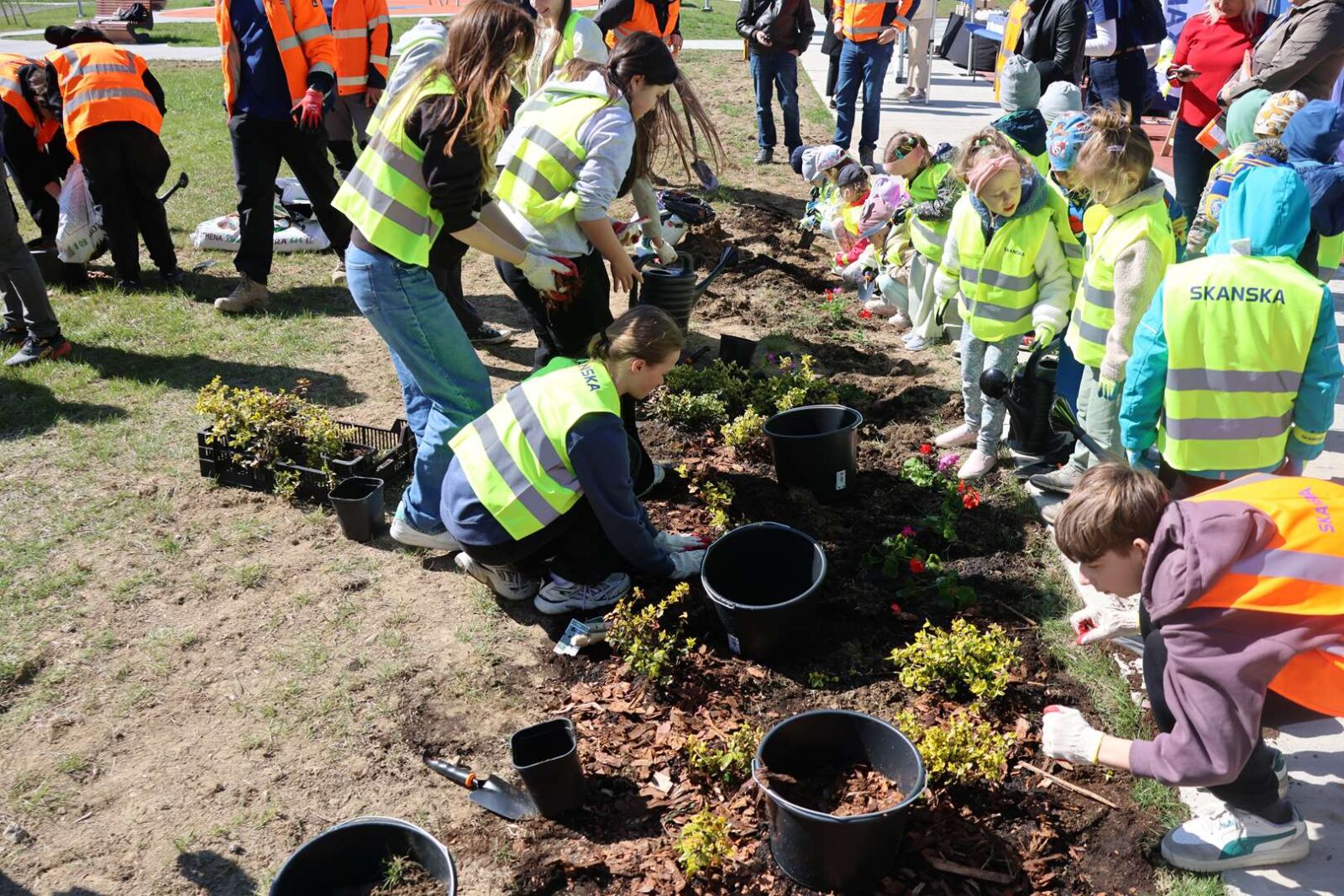 Group of kids in neon safety vests planting flowers in a garden bed under adult supervision outdoors.",