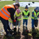 Children in neon safety vests plant a small shrub while an adult helper pours soil from a bucket in a park.