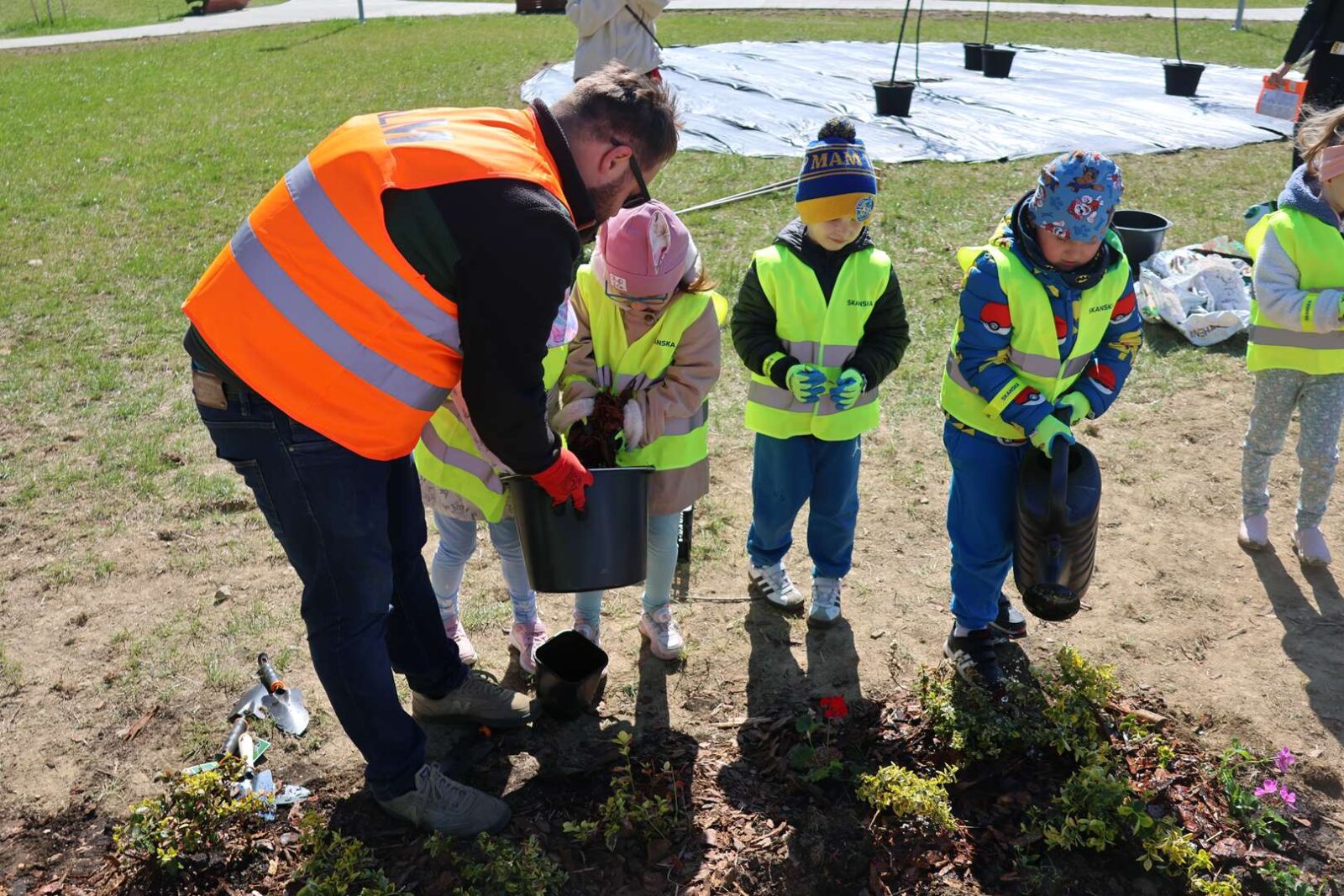 Children in neon safety vests plant a small shrub while an adult helper pours soil from a bucket in a park.