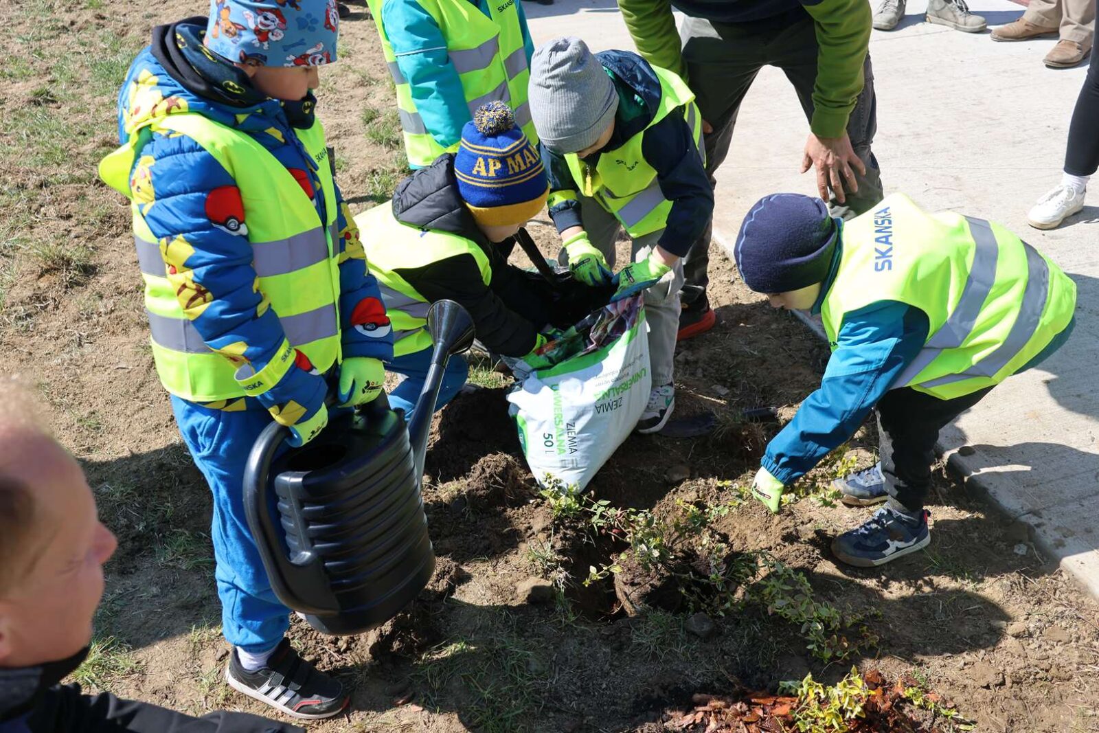 Children in neon safety vests plant a small sapling, filling a dug hole with soil from a bag as part of a outdoor cleanup event.