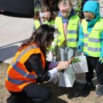 Adult woman in an orange safety vest kneels on the ground handing gift bags to a line of children in bright yellow-green vests outside.