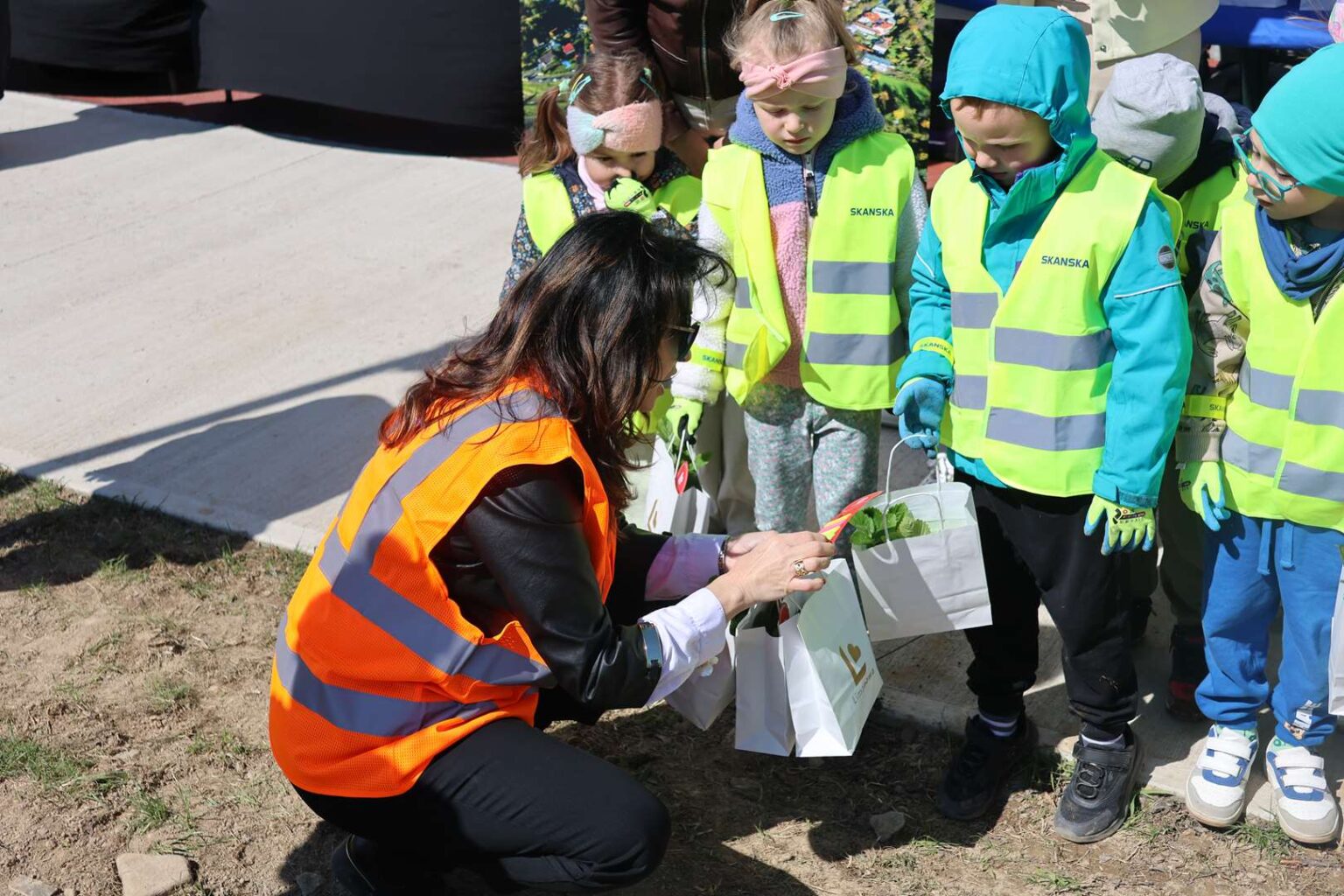 Adult woman in an orange safety vest kneels on the ground handing gift bags to a line of children in bright yellow-green vests outside.