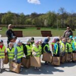 Group of smiling children in neon vests with brown paper bags in a park, supervised by adults against a sunny backdrop.