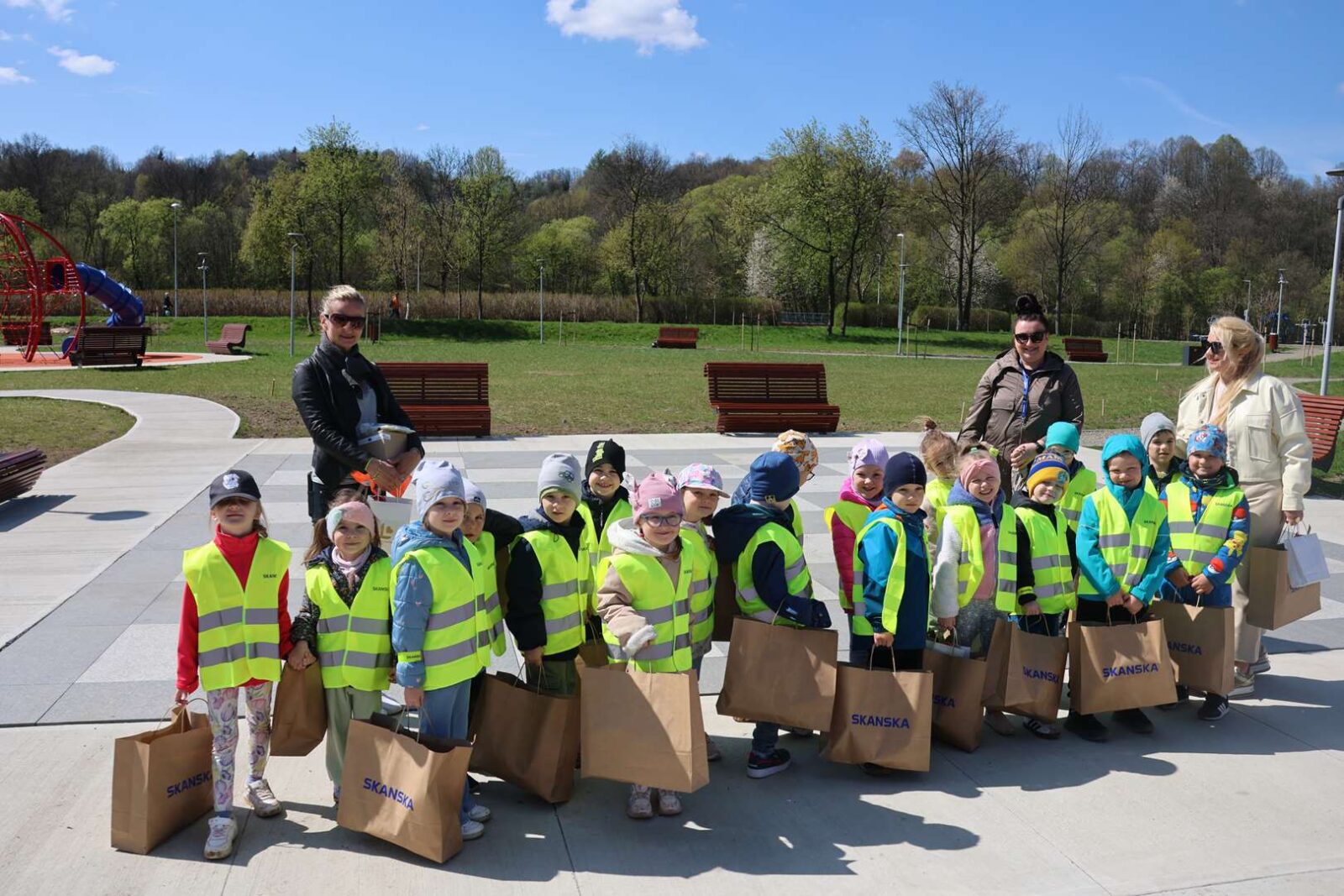 Group of smiling children in neon vests with brown paper bags in a park, supervised by adults against a sunny backdrop.